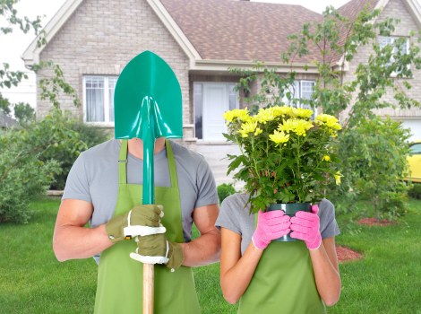 This image features two individuals standing on a well-maintained front lawn in a suburban residential garden. One person is wearing a green apron, a grey t-shirt, and brown gardening gloves, holding a green shovel with a turquoise handle, positioned vertically in front of their torso. The other person, also wearing a grey t-shirt and pink gardening gloves, is holding a large pink pot filled with bright yellow flowers, partially obscuring their face. The background shows a neatly mown green lawn bordered by garden beds with shrubs and young trees. Behind these, there is a modern house with a grey tiled roof, light-colored brick walls, and white-framed windows. The scene appears to be on a sunny day, with natural light illuminating the garden and outdoor environment, emphasizing the active gardening setting. This setup aligns with outdoor landscaping or gardening services offered by Hedge Trimming Barnet, demonstrating maintenance and planting activities typical of professional gardening work in the Barnet area.