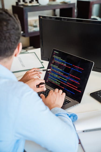 A person working at a desk with two computer monitors, one displaying lines of colorful code, and the other turned off or showing a blank screen. The individual is using a laptop with a black keyboard