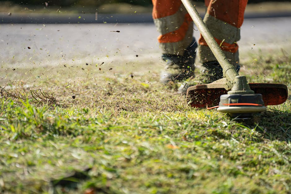 A gardener wearing orange work trousers and grey shoes is using a petrol-powered brush cutter to trim grass and weeds along a garden pathway. The tool has a long handle and a rotating cutting head, wh